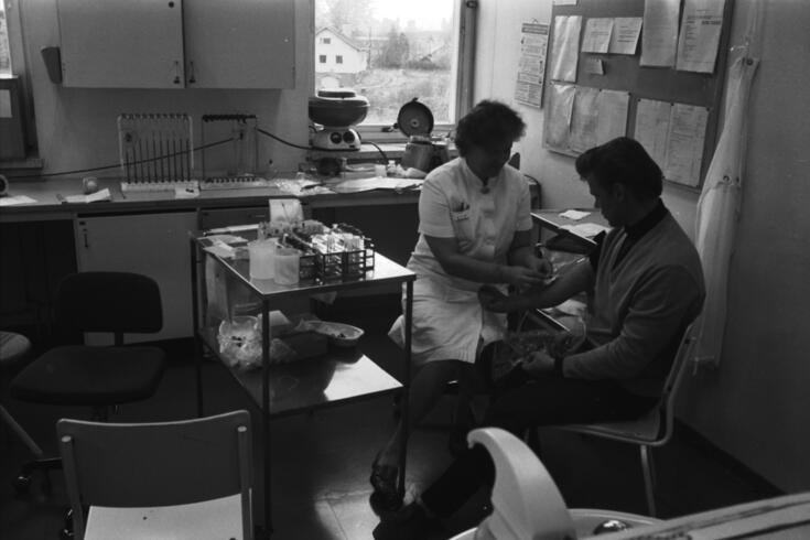 Hoitaja ottaa potilaan käsivarresta verinäytteen mustavalkoisessa valokuvassa. 