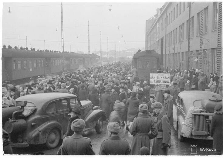 Ihmisjoukko liikkuu Helsingin rautatieasemalla. Kuvassa näkyy kaksi junaa ja etualalla kaksi henkilöautoa. Toisen junan edessä on kyltti, jossa lukee pikajuna Äänislinnaan - Till Äänislinna