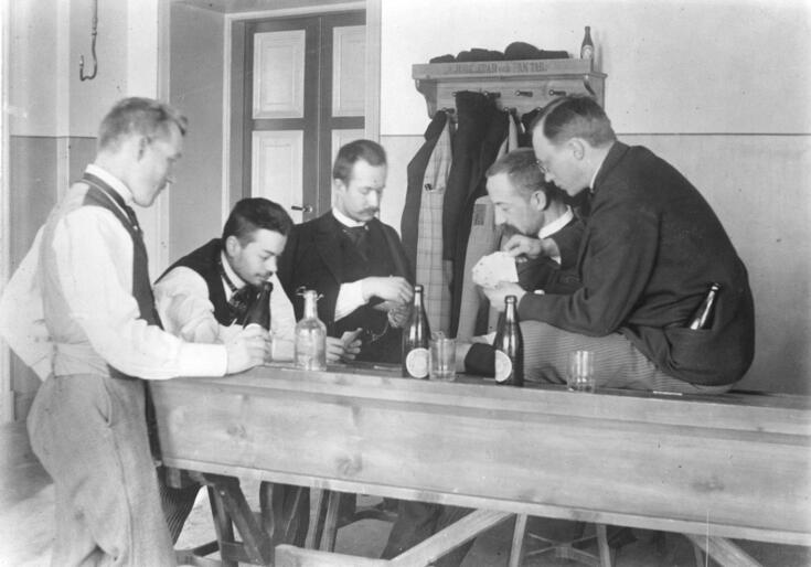 Fem unga män spelar kort samlade vid en arbetsbänk. 