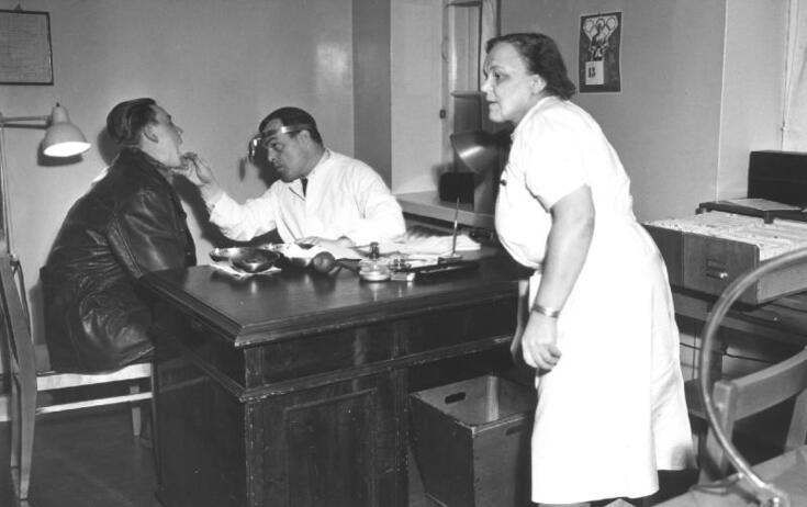 Vasemmalla istuu mies tuolilla suu auki. Keskellä on lääkäri, joka katsoo hänen suuhunsa. Oikealla on sairaanhoitaja. 