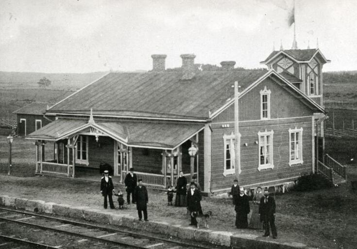 Puolitoistakerroksinen puinen asemarakennus, jonka edessä seisoo ihmisiä.