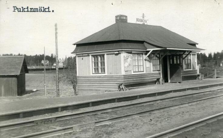 En liten stationsbyggnad i trä i en våning vid järnvägsspåret. På taket finns en skylt med texten BOXBACKA.