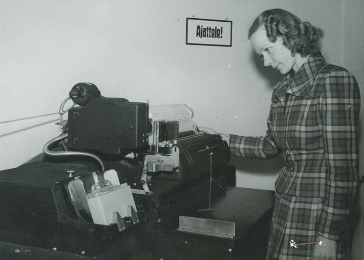 En kvinna i en åtsittande rutig dräkt står vid ett bord och övervakar en maskin som behandlar information.