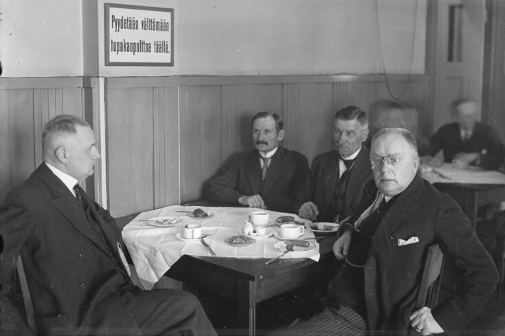 Neljä muodollisesti pukeutunutta miestä istuu valkoisella liinalla peitetyn kahvipöydän ääressä. Seinällä on kyltti: Pyydetään välttämään tupakanpolttoa täällä.