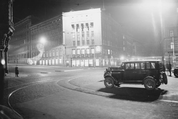 Raken talo Bulevardin ja Erottajan kulmauksessa yövalaistuksessa. Vasemmalla on jalankulkija, etualalla on auto.