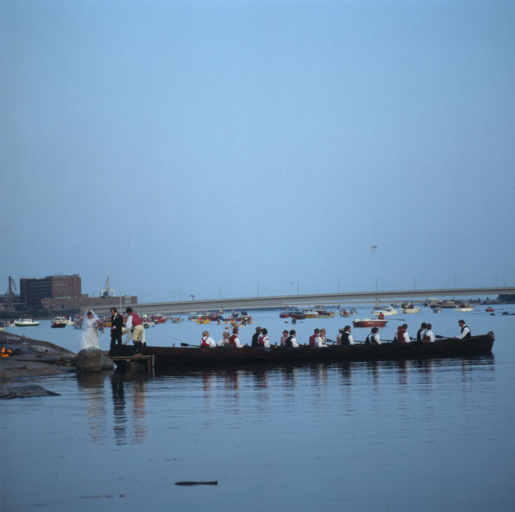 Kirkkovene on soudettu Seurasaaren rantaan, jossa hääpari on nousemassa maihin. Taustalla näkyy Lapinlahden silta ja sen vasemmalla puolen tehdasrakennus.