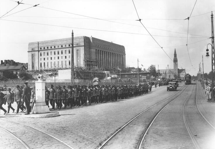 Etualalla näkyy raitiotiekiskoja ja metallinen liikennemajakka. Sen takana marssii iso väkijoukko. Taustalla kohoaa eduskuntatalo, jonka rakentaminen on loppusuoralla.