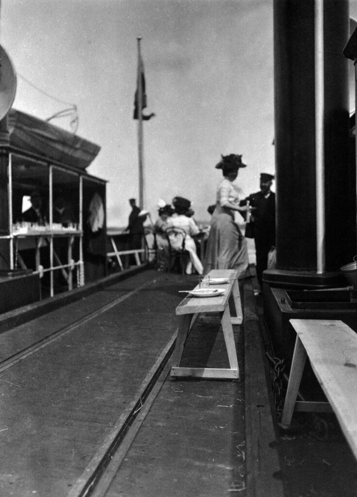 Till vänster syns en spårvagn med öppna väggar. I den står två män i hattar. I mitten syns stolar och små bord kring vilka människor sitter och äter. I förgrunden syns en bänk av trä och på den två använda fat och glas. Bänken omges av spårvägsspår. Två tjänstemän iförda uniformsmössa är också närvarande. 