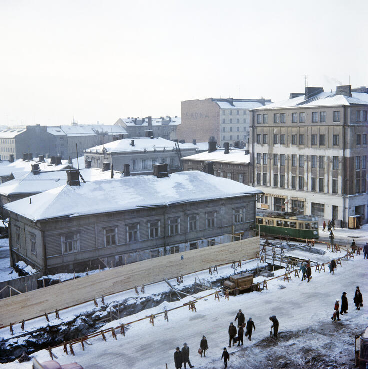 Ylhäällä otetussa kuvassa matalan puutalon edustalla on aita ja kaivanto. Maassa on lunta. Kadulla on jalankulkijoita ja raitiovaunu. Taustalla näkyy sekä puutaloja että kivitaloja. 