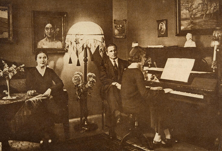 Nainen istuu sohvalla, mies nojatuolissa ja tytär pianon ääressä.