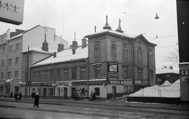 Etualalla on loskaista katua ja jalankulkijoita. Keskellä kolmekerroksinen puurakennus, jonka seinässä on liikekilpiä.