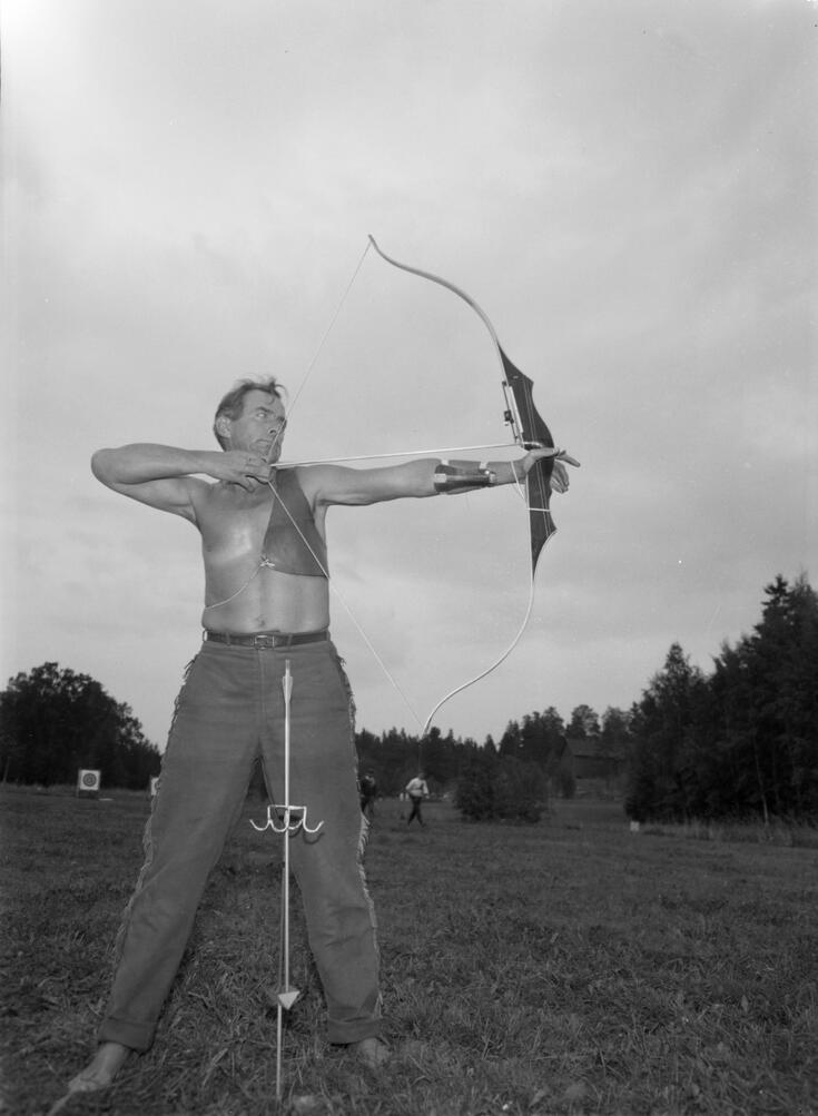 I förgrunden står en barbröstad man som spänner en pilbåge med en pil. I bakgrunden syns måltavlor och andra personer.