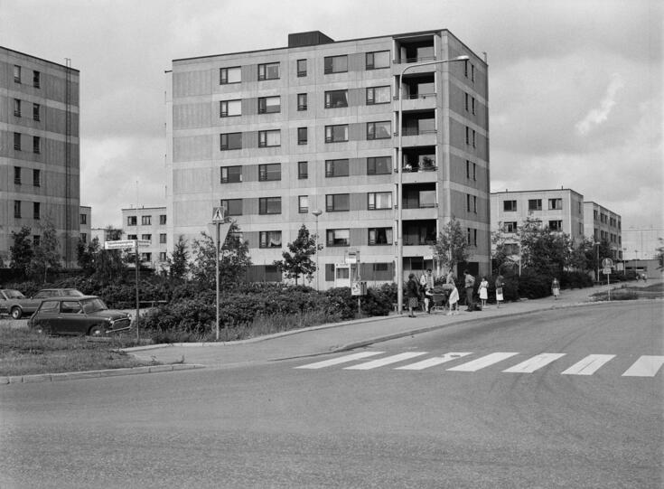 Keskellä on 7-kerroksinen elementtikerrostalo, jonka vieressä on linja-autojen päätepysäkki Ajurinaukiolla. Etualalla on suojatie, vasemmalla on autoja talojen parkkipaikalla. Kuvassa on myös matalampia kerrostaloja.