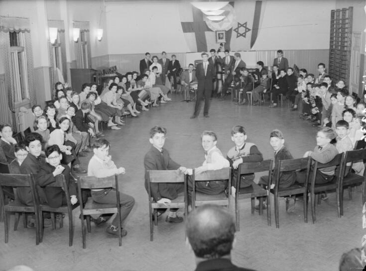 Barn sitter på stolar, som är utplacerade för att bilda en ring. Vuxna står. På den bakre väggen hänger Finlands och Israels flaggor.