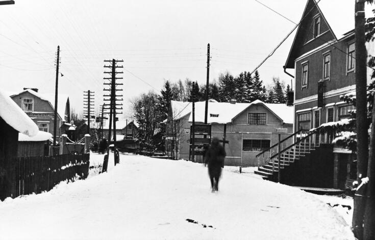 Luminen kylänraitti, jota reunustavat puutalot ja sähköpylväät. Keskellä kävelee yksi ihminen.