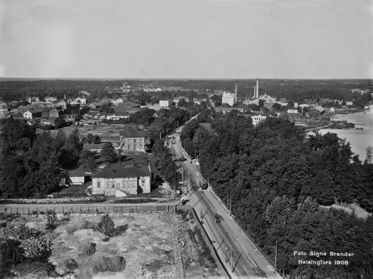 Panoraama Kansallismuseon tornista pohjoiseen. Kuvassa näkyy matalia taloja, puita, kalliota ja maantie.