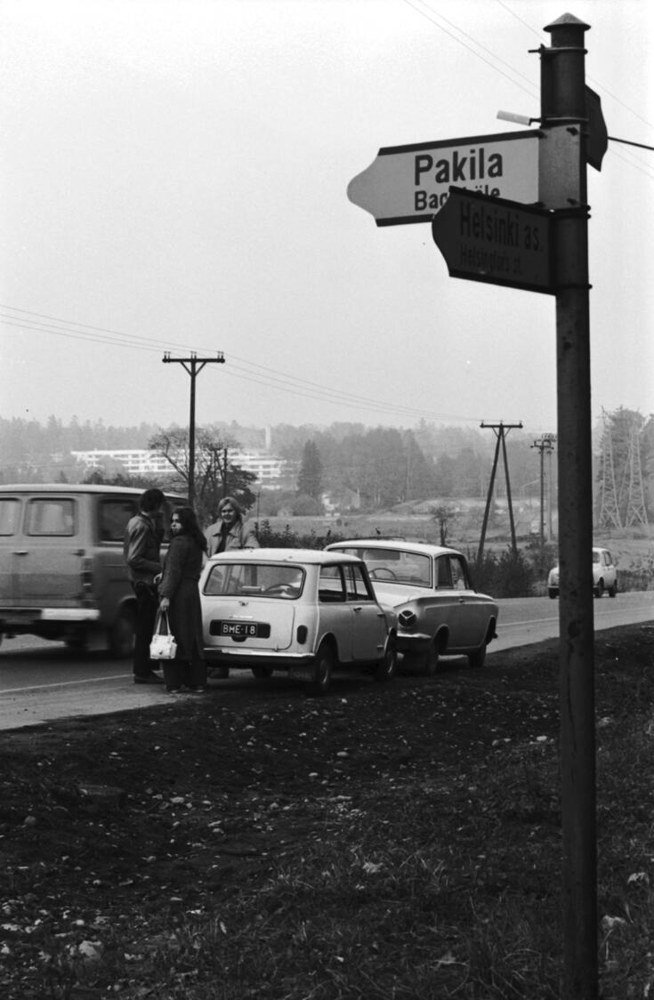 Autoja ja ihmisiä Käskynhaltijantiellä Mikkolantien risteyksessä Oulunkylässä. Etualalla tienviitta Pakilaan. Taustalla Pukinmäen kerrostaloja Sunilantien varrella.