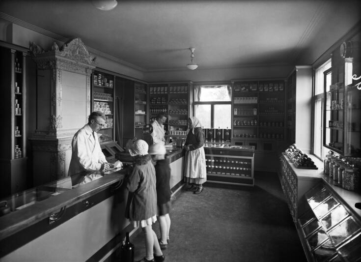 Vasemmalla on pitkä myyntitiski, jonka takana seisoo kaksi valkoiseen takkiin pukeutunutta myyjää. Toinen myyjä palvelee huivipäistä naista ja toinen kahta lasta. Myymälän seiniä reunustavat tuotehyllyt. Myyjien takana on nelikulmainen kaakeliuuni.