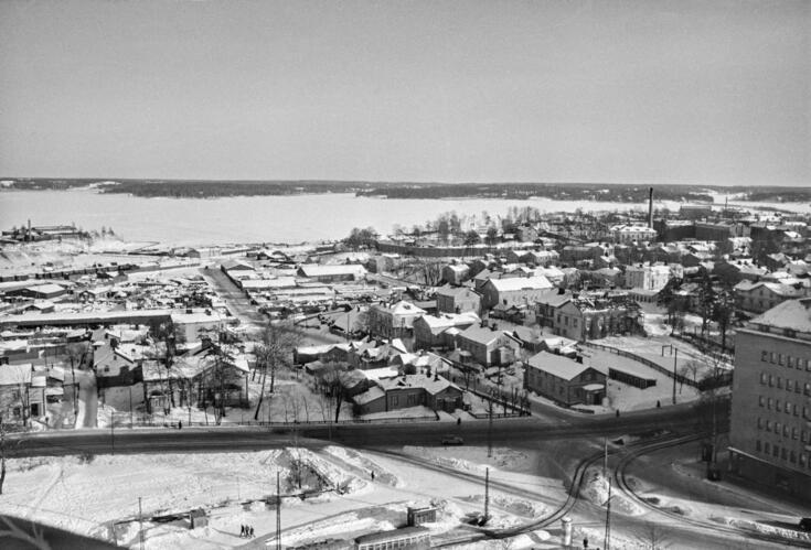 Katoilla ja maassa on lunta. Etualalla raitiovaunu kaartaa kohti kirkkoa kiertävää silmukkaa, oikealla on Sturenkadun risteys. Hämeentien toisella puolella näkyy Hermannin matalia puutaloja, niiden takana viljelypalstoja ja oikealla tiilimuurin aitaama vankila-alue. Vasemmalla Kyläsaaren jätteenpolttolaitoksen edestä kulkee tavarajuna. Rantaa on vuosikymmenten kuluessa täytetty, ja maa-alue on lisääntynyt melkoisesti. Vanhankaupunginlahden takana näkyvät oikealta Kulosaari, Kivinokka ja Herttoniemi.