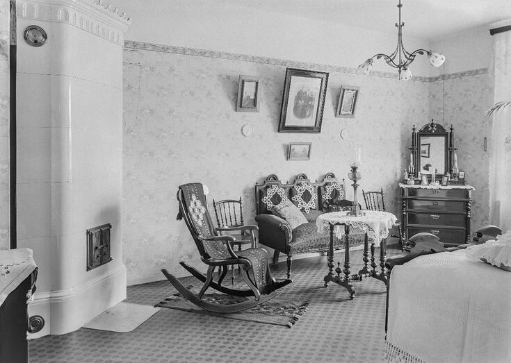 Det ljusa rummet har an vit kakelugn till vänster, mönstrade tapeter och tavlor på väggarna, en gungstol, soffa och säng, ett dekorerat lillbord samt en dekorativ spegelbyrå. Det är snyggt och hemtrevligt.