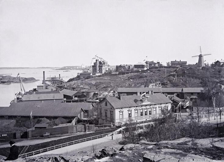 Etualalla kalliota, satamarata ja Saunakatu. Satamarakennuksia ja laivatelakka, tuulimylly ja taustalla merta.