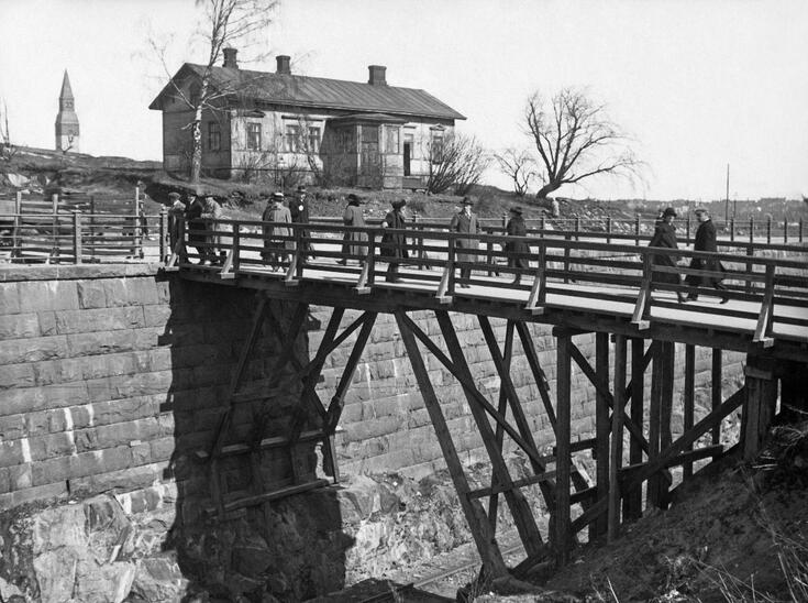 Ihmisiä Arkadiankadun junaradan yli kulkevalla puusillalla. Kuvan vasemmassa reunassa näkyy Kansallismuseon torni. Sillan toisella puolella on yksikerroksinen puutalo sekä muutama paljas koivu.