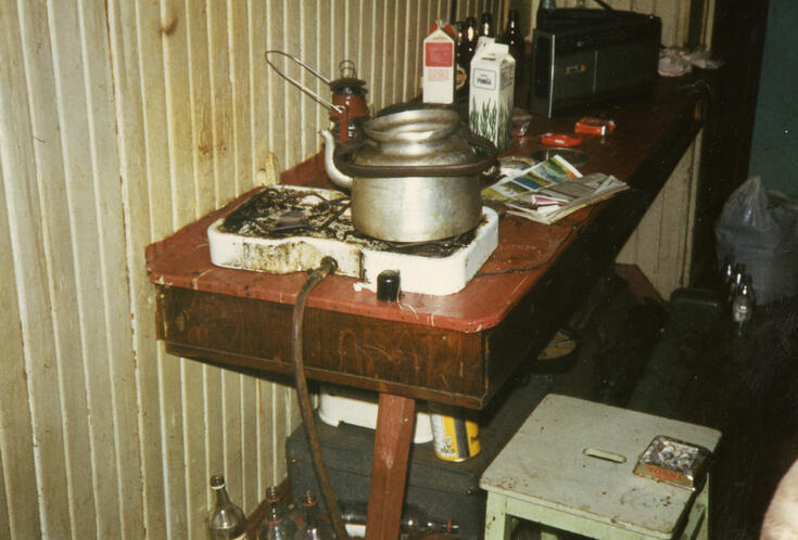 A cramped space with a hotplate and an aluminium coffee pot on a small table. There are food packages on the table, empty bottles on the floor and stains on the wall.