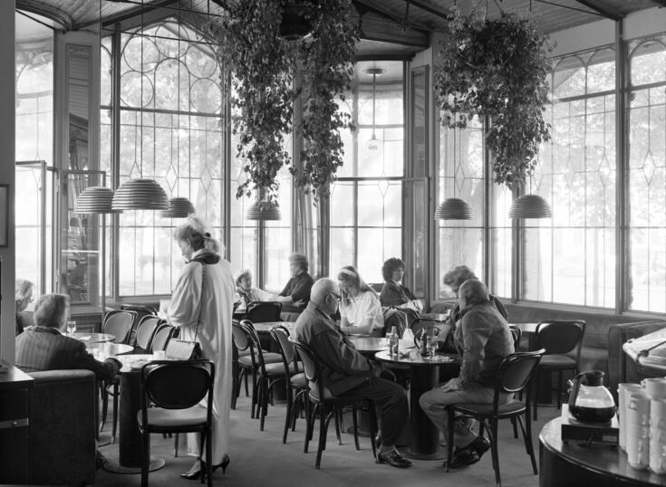 Restaurang Kappelis kafésida. Kunder sitter vid avlånga bord. En kvinna år på väg att sätta sig. 