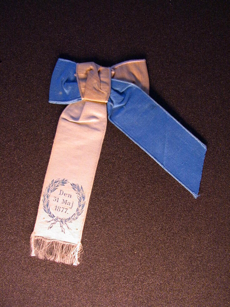 Blåvit rosett, det vita bandet är längre, i änden finns fransar. Datumet och lagerbladskransen har tryckts med blått.