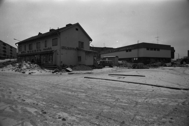 Till vänster en äldre byggnad med ljusreklamen Elanto, baktill till höger en nyare byggnad med ljusreklamen Alennushalli Elanto Lågprishall. Framför den gamla byggnaden finns en stor snöhög, och det ligger byggavfall runt båda byggnaderna.