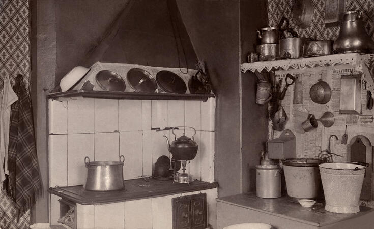 Vasemmalla on puuhella, oikealla on senkki, jonka päällä on sankoja. Seinällä on keittiövälineitä ja seinällä kattilahylly.