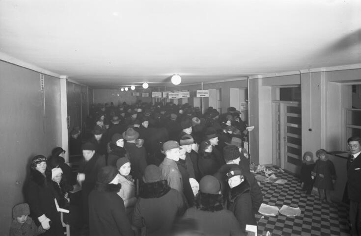 Pitkänomainen matalahko tila, jossa tungeksii ulkovaatteisiin pukeutuneita ihmisiä paperipinoja käsissään. He jonottavat luukuille, joiden yläpuolella katosta roikkuu kyltit. Kuvan oikeassa reunassa seisoo kaksi lasta ja vahtimestari oven vieressä.