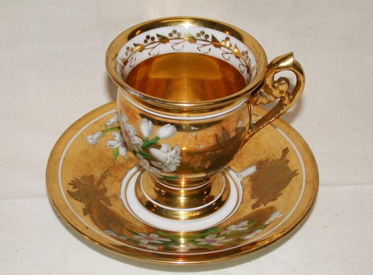 Siro kahvikuppi ja lautanen, jotka on kullattu ja koristeltu valkoisin narsissein.