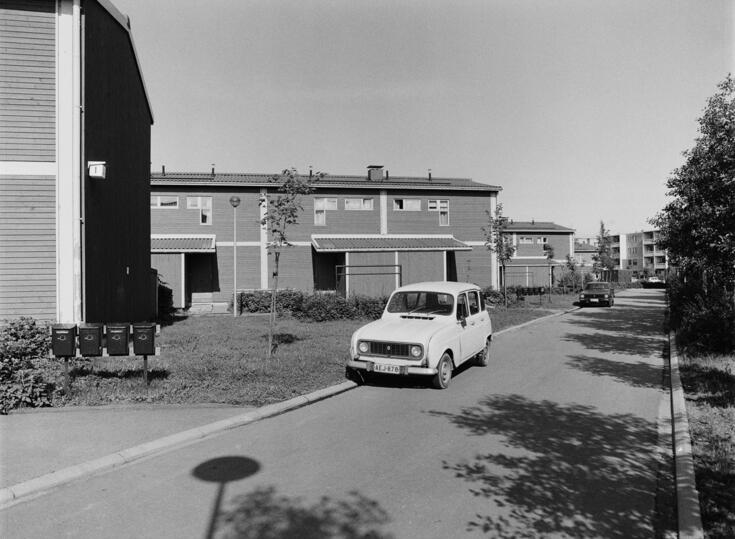 Vasemmalla on kaksikerroksisia rivitaloja, etualalla on tie, jonka varrella on kaksi autoa. Oikeassa reunassa on puita, ja taustalla kohoaa nelikerroksinen kerrostalo.