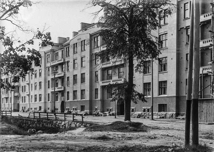 Kerrostalo, jossa on neljä asuinkerrosta. Talon edessä kasvaa muutama puu.