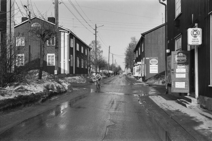 Katunäkymä Limingantieltä Jyrängöntien kulmasta koilliseen kevätauringon sulattaessa lumia. Oikealla kaksikerroksisissa puutaloissa myymälöitä, edessä Limingantie 9:ssä H.M. Laineen K-kauppa.
