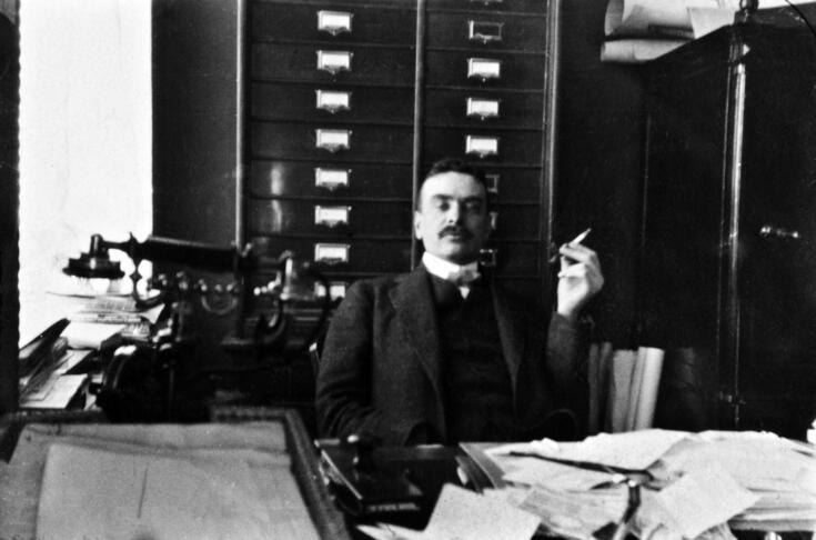 En man med mustasch sitter bakom ett skrivbord med en cigarett i handen. Bakom honom syns ett arkivskåp och ett kassaskåp. I förgrunden på arbetsbordet finns mycket papper och en telefon.