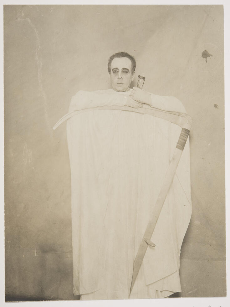 Valkoiseen huputtomaan kaapuun pukeutunut voimakkaasti maskeerattu mies seisoo nojaamassa viikatteen terään.