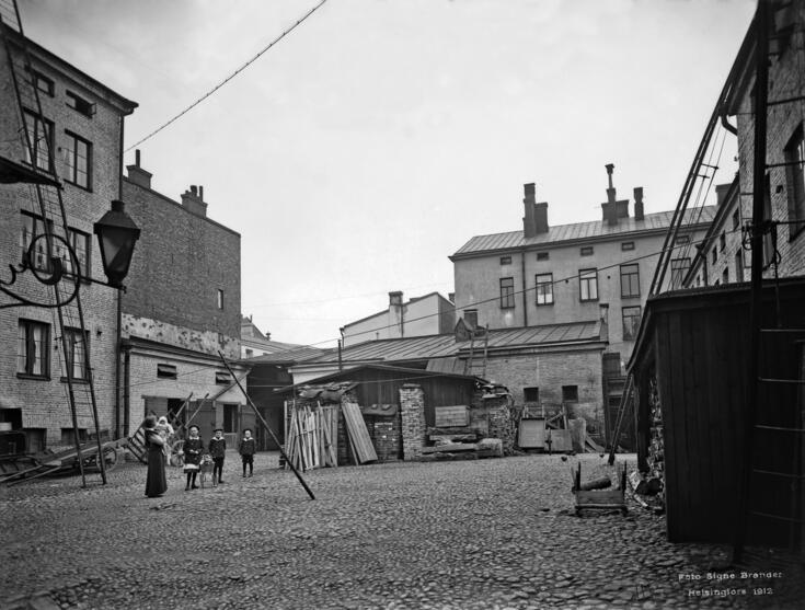 Aleksanterinkatu 52, sisäpiha. Sivuttain katsojaan seisoo nainen, jolla on lapsi sylissä. Kolme isompaa lasta seisoo rivissä keskellä pihaa. 