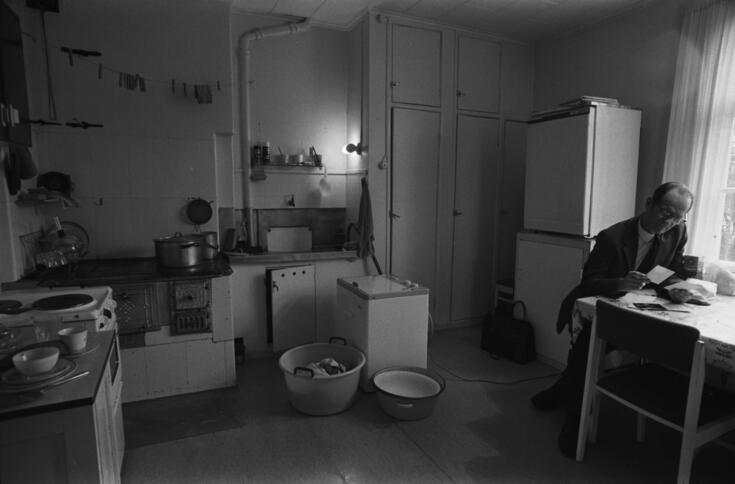 Keittiö sähkö- ja puuliesineen sekä pyykinpesuvälineineen, mies pöydän ääressä Puu-Käpylässä ennen peruskorjausta. 