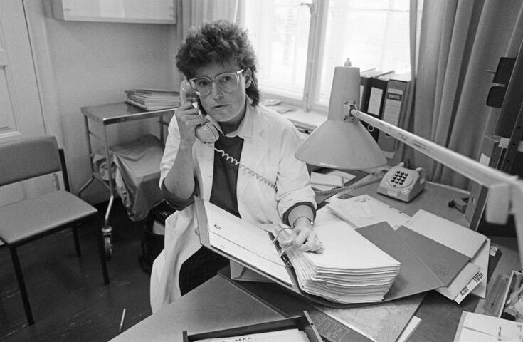 Lääkärintakkiin pukeutunut silmälasipäinen nainen istuu kirjoituspöydän takana puhelimen kuuloke korvallaan. Hänen edessään on avoimena paksu mappi.