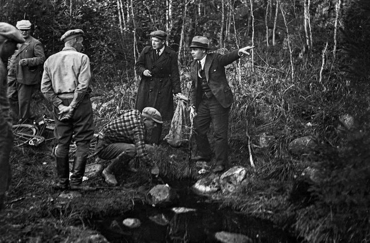 Keskellä kuvaa on ruudulliseen paitaan ja lippalakkiin pukeutunut mies kumartuneena lähteen ylle. Oikealla seisoo pukuun ja hattuun pukeutunut nimismies, joka osoittaa vasemmalla kädellään poispäin. Kuvassa on myös neljä muuta miestä ja maassa makaava polkupyörä.