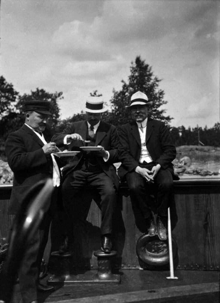 Tre män på däcket till spårvagnsångfärjan. Mannen till vänster står och äter, mannen i mitten sitter på relingen och äter, och mannen till höger sitter på relingen med händerna i knät och ler mot kameran.