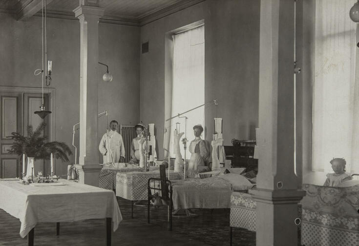 På bilden är två sjuksköterskor, en läkare och fyra barnpatienter, som alla har en nedre extremitet i en draganordning. I bildens högra kant är en flicka som besöker sjukhuset.