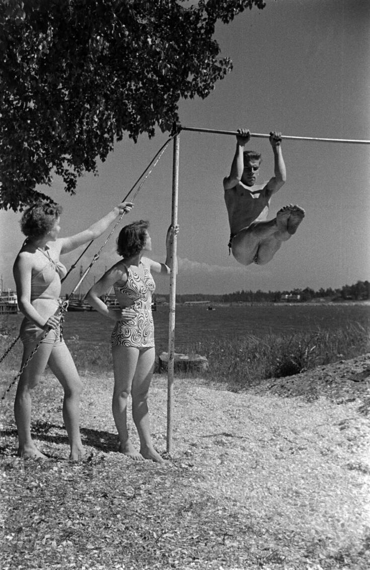 Kaksi naista ja mies Sompasaaren rannalla. Mies voimistelee rekkitangolla.