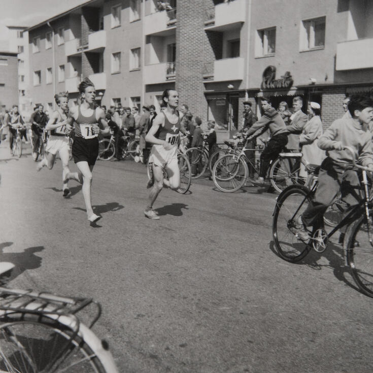 Längs vägen springer människor med nummerlapp på bröstet. Bakom dem syns cyklister.