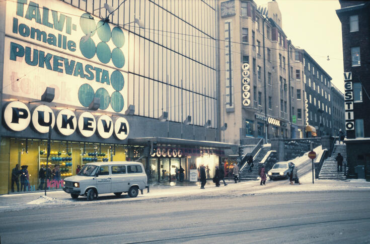 Kajsaniemigatan 9, 5, Berggatan 7. Till höger Berggatan 10. Pukevas varuhus till vänster, Vestios klädbutik till höger. Bredvid Vestio en trappa till Berggatan. På Pukevas yttervägg hänger ett reklamplakat med texten: "Talvilomalle Pukevasta". Till höger Vestios ljusreklam.