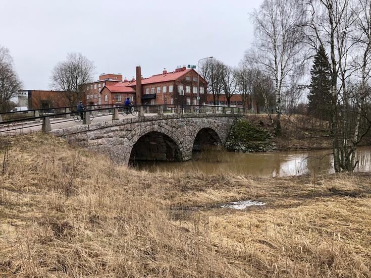 Brobackan vanha kivisilta Helsingin ja Vantaan rajalla. Rajakyltti on sillan keskikohdassa kaiteen luona. SIllan ali virtaa Kerava-joki. Taustalla näkyvät tehdasrakennukset ovat osoitteessa Kirkonkyläntie 103. Sillalla kulkee kaksi polkupyöräilijää.