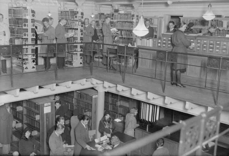 Näkymä Richardinkadun kirjaston kahteen kerrokseen. Ylemmässä kerroksessa asiakkaat tutkivat kirjoja ja kortistoa, alemmassa jonotetaan palvelutiskille.