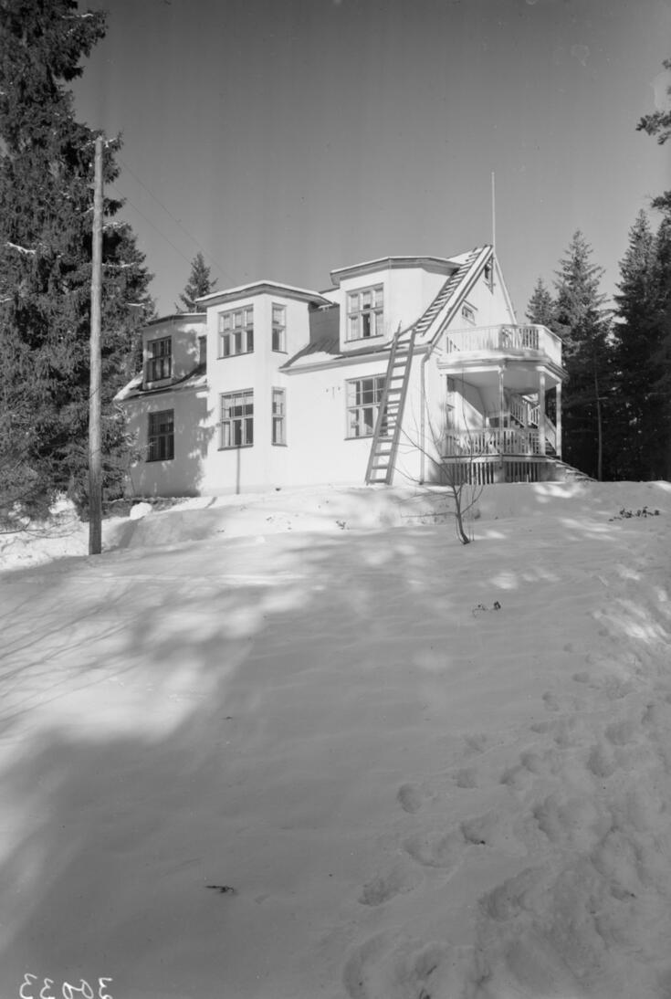 En vitmålad trävilla i två våningar, som har en veranda på kortsidan och ovanför den en balkong. Det är mycket snö runt huset.